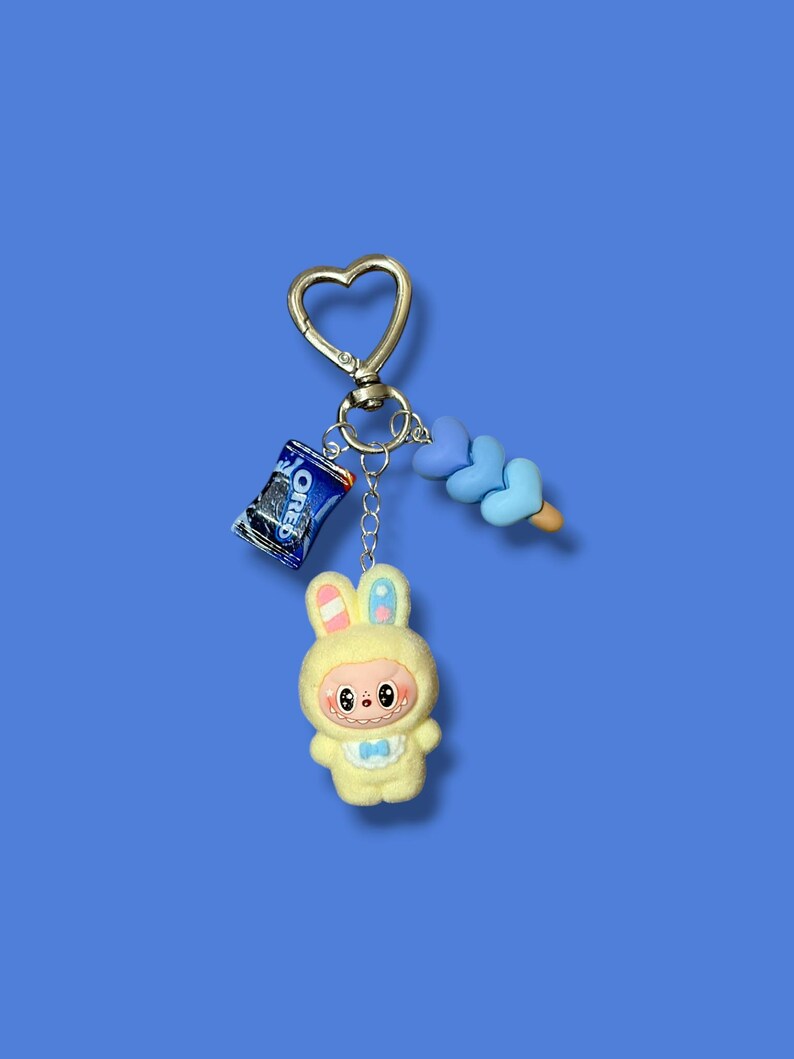 May include: A whimsical keychain featuring a yellow bunny character in a hooded outfit, a miniature Oreo cookie package, and a blue heart-shaped candy. The keychain has a gold-colored heart-shaped clasp and a blue background.