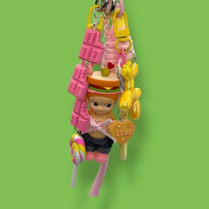 May include: A whimsical keychain featuring a variety of charms. Includes a figure in denim shorts, a sandwich, pink chocolate bars, a lollipop, a balloon dog, and a heart-shaped charm with the word "LOVE". The keychain is attached to a silver chain.