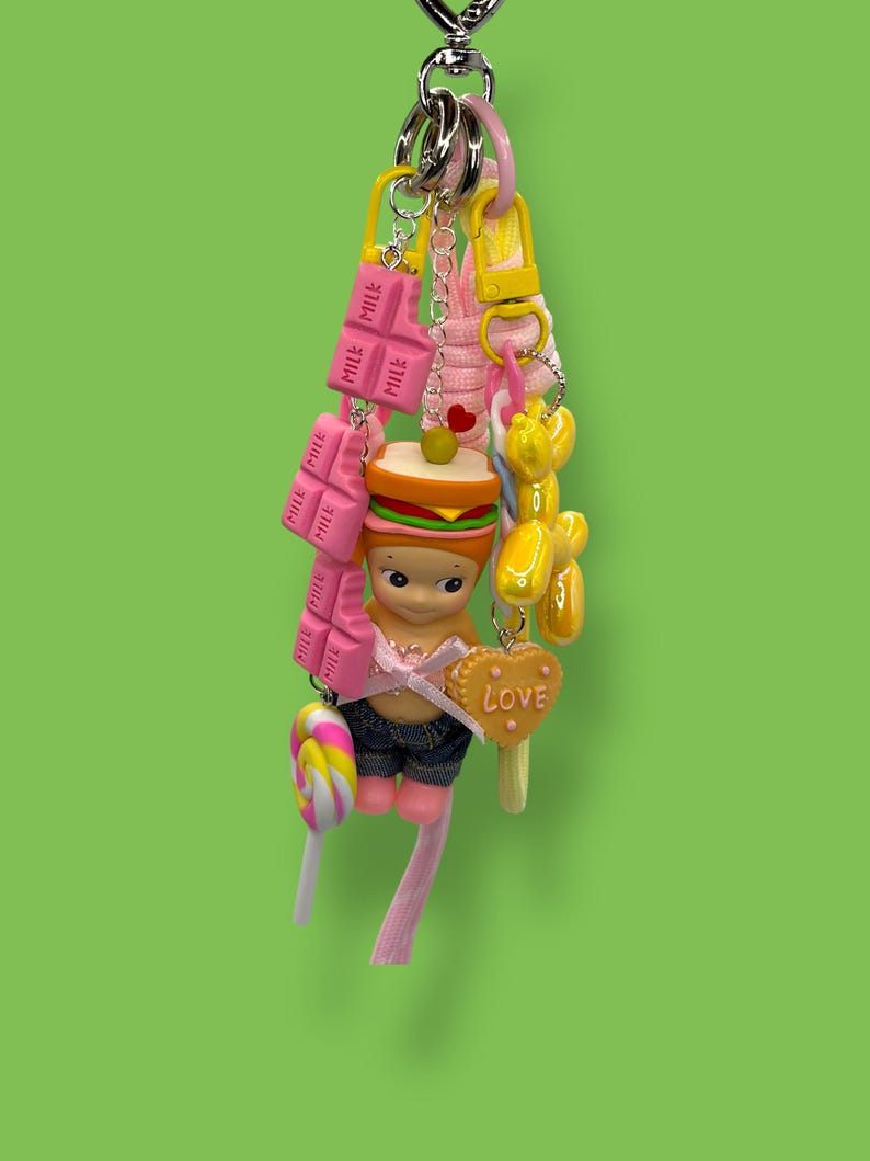 May include: A whimsical keychain featuring a variety of charms. Includes a figure in denim shorts, a sandwich, pink chocolate bars, a lollipop, a balloon dog, and a heart-shaped charm with the word "LOVE". The keychain is attached to a silver chain.