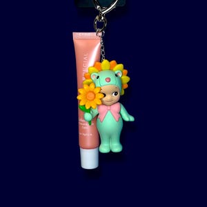 K&ouml;nnte beinhalten: Ein Schl&uuml;sselanh&auml;nger mit einer gr&uuml;nen Figur, die einen Sonnenblumenhut und eine rosa Schleife tr&auml;gt und eine gelbe Blume h&auml;lt. Eine Tube Lippenbalsam ist befestigt. Der Schl&uuml;sselanh&auml;nger hat einen silbernen herzf&ouml;rmigen Verschluss.