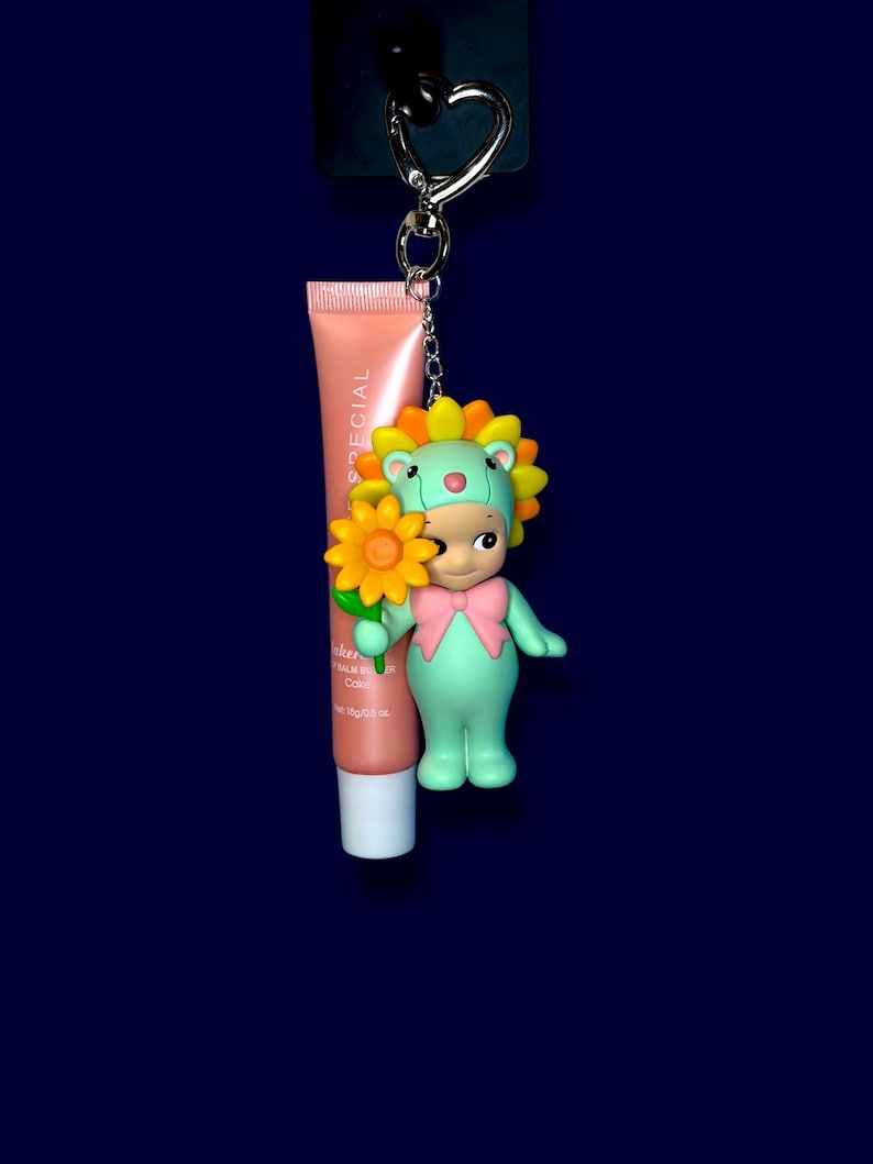 K&ouml;nnte beinhalten: Ein Schl&uuml;sselanh&auml;nger mit einer gr&uuml;nen Figur, die einen Sonnenblumenhut und eine rosa Schleife tr&auml;gt und eine gelbe Blume h&auml;lt. Eine Tube Lippenbalsam ist befestigt. Der Schl&uuml;sselanh&auml;nger hat einen silbernen herzf&ouml;rmigen Verschluss.