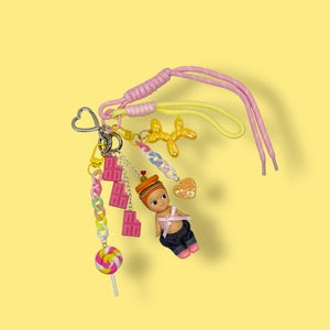May include: A colorful keychain with multiple charms, including a small doll, a lollipop, and a gold-colored balloon dog. The keychain features a pink and yellow shoelace-style strap and a heart-shaped clasp. The background is a solid yellow.