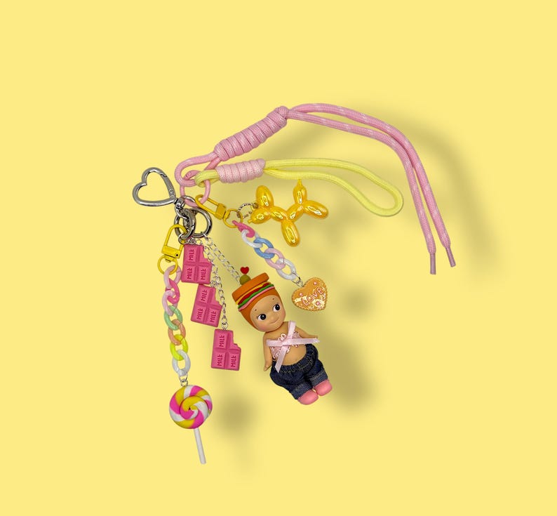 May include: A colorful keychain with multiple charms, including a small doll, a lollipop, and a gold-colored balloon dog. The keychain features a pink and yellow shoelace-style strap and a heart-shaped clasp. The background is a solid yellow.