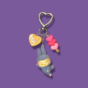 May include: A silver heart-shaped keychain with three charms: a yellow heart-shaped cookie with the word "LOVE", a blue bunny figure, and a pink heart-shaped popsicle. The charms hang against a purple background.