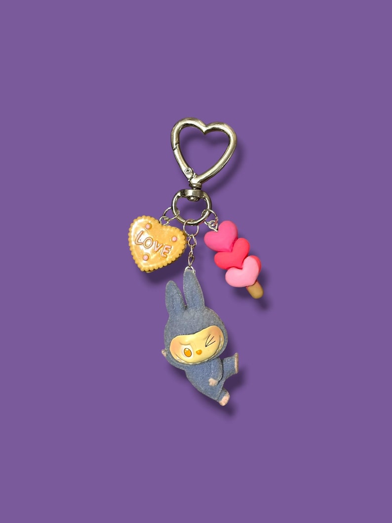 May include: A silver heart-shaped keychain with three charms: a yellow heart-shaped cookie with the word "LOVE", a blue bunny figure, and a pink heart-shaped popsicle. The charms hang against a purple background.