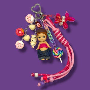 May include: A vibrant keychain featuring a doll figure, miniature donuts, hearts, and a balloon dog charm. The keychain includes a pink and white braided lanyard, a rainbow lollipop, and a heart-shaped clasp. The background is a solid purple.