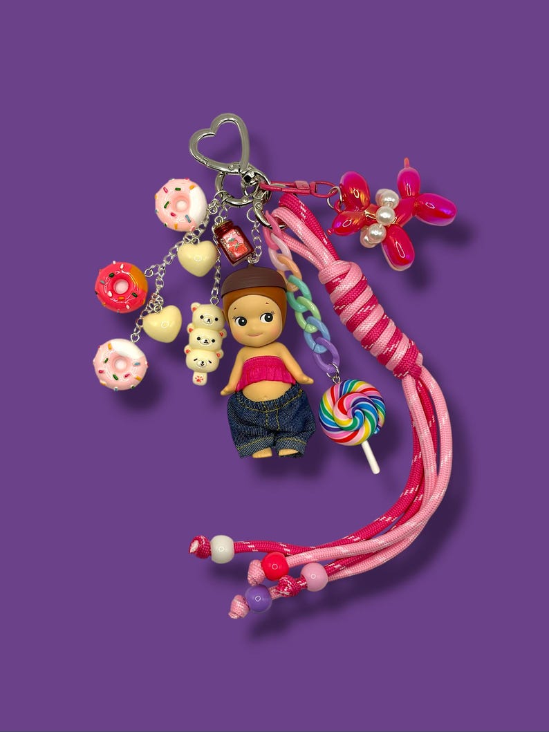 May include: A vibrant keychain featuring a doll figure, miniature donuts, hearts, and a balloon dog charm. The keychain includes a pink and white braided lanyard, a rainbow lollipop, and a heart-shaped clasp. The background is a solid purple.