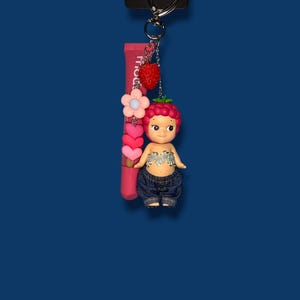 May include: A keychain with a strawberry-themed doll, pink lip gloss, and a strawberry charm. The doll has a berry-like head, denim shorts, and a silver top. The lip gloss tube is pink with white text.