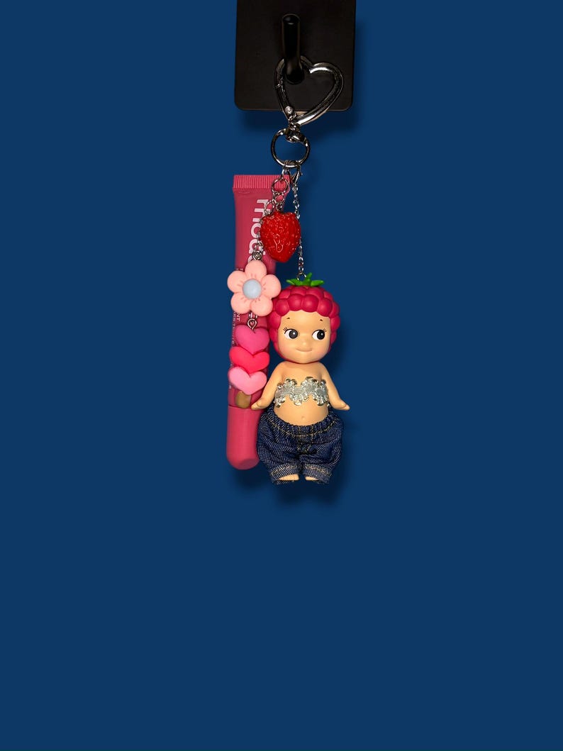 May include: A keychain with a strawberry-themed doll, pink lip gloss, and a strawberry charm. The doll has a berry-like head, denim shorts, and a silver top. The lip gloss tube is pink with white text.