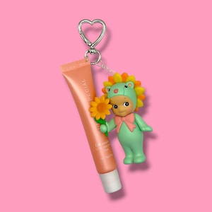 K&ouml;nnte beinhalten: Ein Schl&uuml;sselanh&auml;nger mit herzf&ouml;rmigem Verschluss, der eine Lippenbalsam-Tube und eine kleine Figur h&auml;lt. Die pfirsichfarbene Lippenbalsam-Tube tr&auml;gt den Aufdruck "SPECIAL". Die Figur ist eine gr&uuml;ne Figur mit Sonnenblumen-Kopfschmuck und einer rosa Schleife.