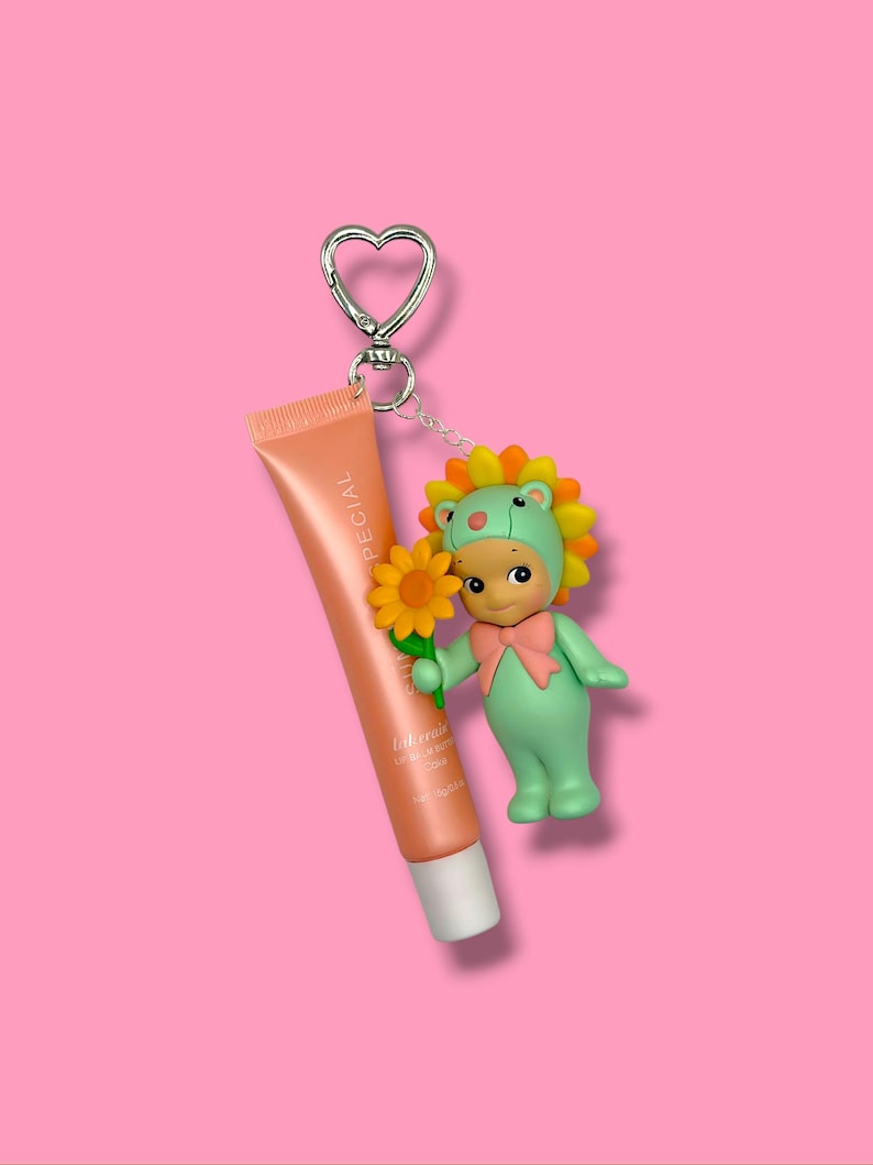 K&ouml;nnte beinhalten: Ein Schl&uuml;sselanh&auml;nger mit herzf&ouml;rmigem Verschluss, der eine Lippenbalsam-Tube und eine kleine Figur h&auml;lt. Die pfirsichfarbene Lippenbalsam-Tube tr&auml;gt den Aufdruck "SPECIAL". Die Figur ist eine gr&uuml;ne Figur mit Sonnenblumen-Kopfschmuck und einer rosa Schleife.