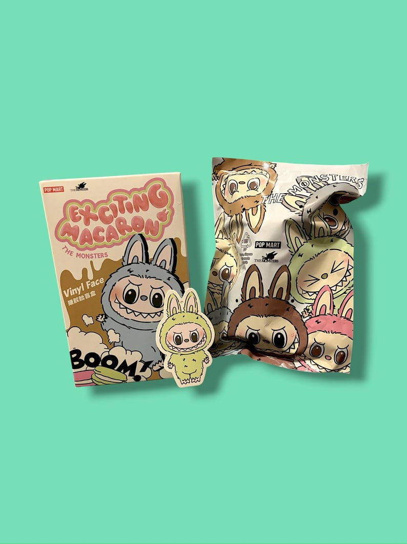 K&ouml;nnte beinhalten: Ein Sammlerspielzeug-Set mit "The Monsters"-Charakteren. Das Set enth&auml;lt eine Box mit dem Text "Exciting Macaron" und einen Blind Bag mit Charakterillustrationen. Ein kleiner Aufkleber eines Charakters ist ebenfalls vorhanden.