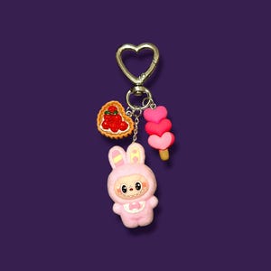 May include: A pink bunny keychain with a heart-shaped clasp. The keychain features a bunny character, a heart-shaped tart with red berries, and a pink heart-shaped popsicle. The background is a solid purple.