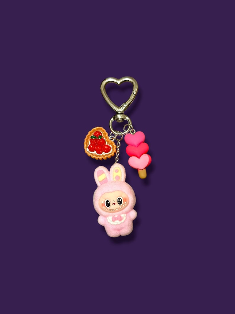 May include: A pink bunny keychain with a heart-shaped clasp. The keychain features a bunny character, a heart-shaped tart with red berries, and a pink heart-shaped popsicle. The background is a solid purple.