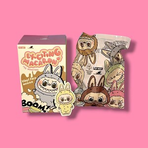 K&ouml;nnte beinhalten: Eine Schachtel und eine T&uuml;te mit "Exciting Macaron The Monsters" Vinyl-Gesichtsfiguren, mit einem Aufkleber. Die Schachtel ist braun und rosa mit Cartoon-Monster-Illustrationen. Die T&uuml;te ist wei&szlig; mit Monster-Illustrationen. Ein gelber Aufkleber eines Monsters ist ebenfalls vorhanden.