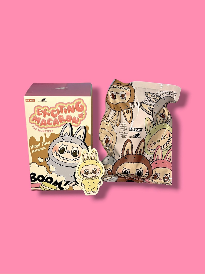 K&ouml;nnte beinhalten: Eine Schachtel und eine T&uuml;te mit "Exciting Macaron The Monsters" Vinyl-Gesichtsfiguren, mit einem Aufkleber. Die Schachtel ist braun und rosa mit Cartoon-Monster-Illustrationen. Die T&uuml;te ist wei&szlig; mit Monster-Illustrationen. Ein gelber Aufkleber eines Monsters ist ebenfalls vorhanden.