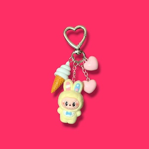 May include: A keychain with a silver heart-shaped clasp, featuring a yellow bunny figure, a white ice cream cone, and two pink hearts. The bunny is wearing a blue bow tie and has blue and pink accents on its ears.