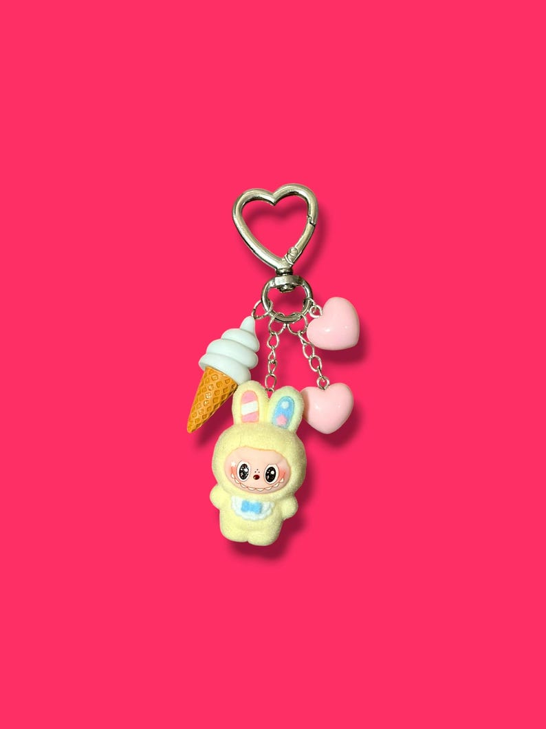 May include: A keychain with a silver heart-shaped clasp, featuring a yellow bunny figure, a white ice cream cone, and two pink hearts. The bunny is wearing a blue bow tie and has blue and pink accents on its ears.