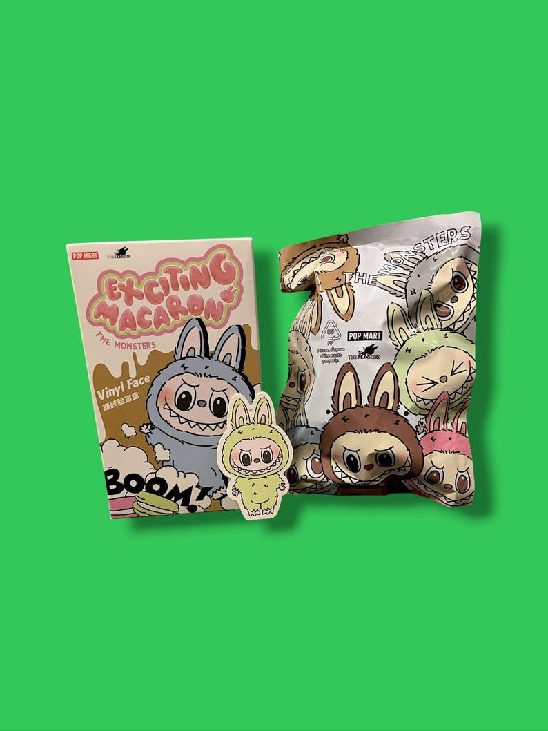 K&ouml;nnte beinhalten: Eine Pop Mart Produktdarstellung mit einer Box und einer T&uuml;te mit Cartoon-Monster-Designs. Auf der Box steht "Exciting Macaron" und "The Monsters". Ein Aufkleber eines Monsters in einem Hasenkost&uuml;m ist ebenfalls vorhanden. Die T&uuml;te ist mit verschiedenen Monsterfiguren gef&uuml;llt.