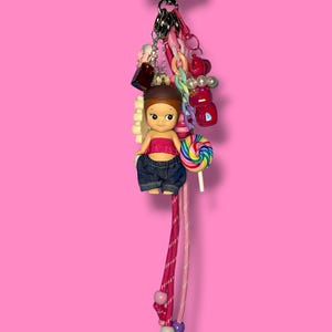 May include: A vibrant keychain with a doll in a pink top and denim jeans, holding a lollipop. The keychain is adorned with charms, beads, and a heart-shaped clasp. The background is a solid pink.