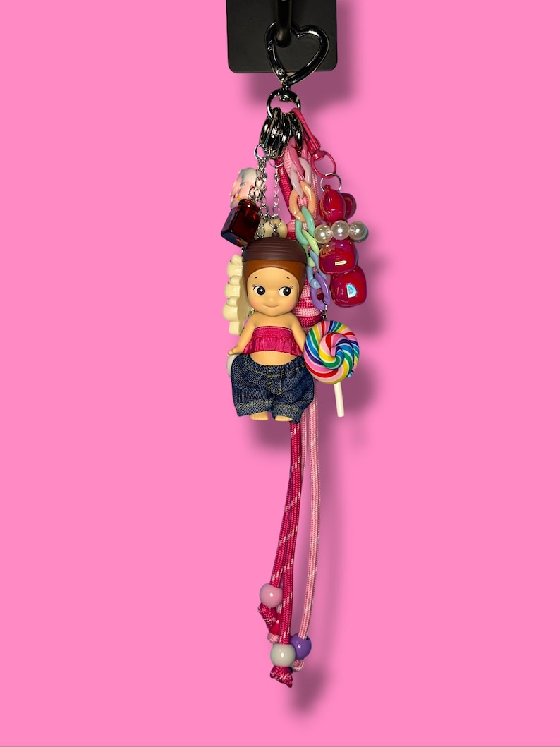 May include: A vibrant keychain with a doll in a pink top and denim jeans, holding a lollipop. The keychain is adorned with charms, beads, and a heart-shaped clasp. The background is a solid pink.