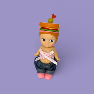 May include: A whimsical figurine with a sandwich-shaped head, featuring layers of bread, lettuce, tomato, and cheese, topped with an olive and a heart-shaped pick. The figurine wears a pink bow top, denim shorts, and pink shoes. The background is a solid purple.