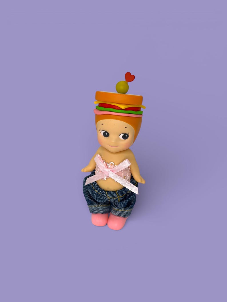 May include: A whimsical figurine with a sandwich-shaped head, featuring layers of bread, lettuce, tomato, and cheese, topped with an olive and a heart-shaped pick. The figurine wears a pink bow top, denim shorts, and pink shoes. The background is a solid purple.