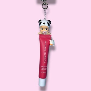 Op de afbeelding: Een sleutelhanger met een rode lippenbalsem tube met de tekst "SUMMER SPECIAL" en een panda-thema babyfiguur. Een kleine koeienbedel hangt aan de tube. De lippenbalsem is gelabeld met "lakerain" en "Pink Guava".