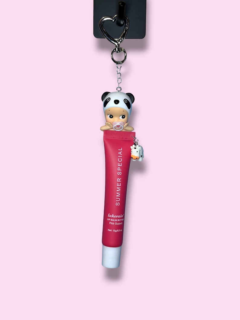 Op de afbeelding: Een sleutelhanger met een rode lippenbalsem tube met de tekst "SUMMER SPECIAL" en een panda-thema babyfiguur. Een kleine koeienbedel hangt aan de tube. De lippenbalsem is gelabeld met "lakerain" en "Pink Guava".