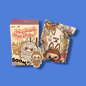 K&ouml;nnte beinhalten: Eine rosa-beige Box mit dem Text "Exciting Macaron The Monsters" und einem Cartoon-Hasencharakter. Ein passender Hasenaufkleber und eine gemusterte Tasche mit Hasencharakteren sind ebenfalls sichtbar. Der Hintergrund ist einfarbig blau.