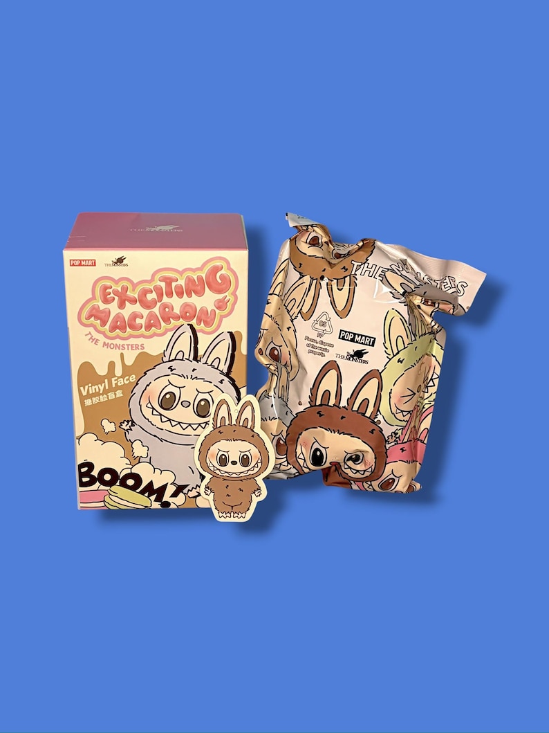 K&ouml;nnte beinhalten: Eine rosa-beige Box mit dem Text "Exciting Macaron The Monsters" und einem Cartoon-Hasencharakter. Ein passender Hasenaufkleber und eine gemusterte Tasche mit Hasencharakteren sind ebenfalls sichtbar. Der Hintergrund ist einfarbig blau.