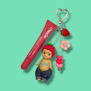 May include: A pink lip tint tube with the brand name "rhode" and a small doll keychain. The doll has a strawberry-shaped head, denim shorts, and a silver top. The keychain also includes a strawberry, flower, and heart charms.