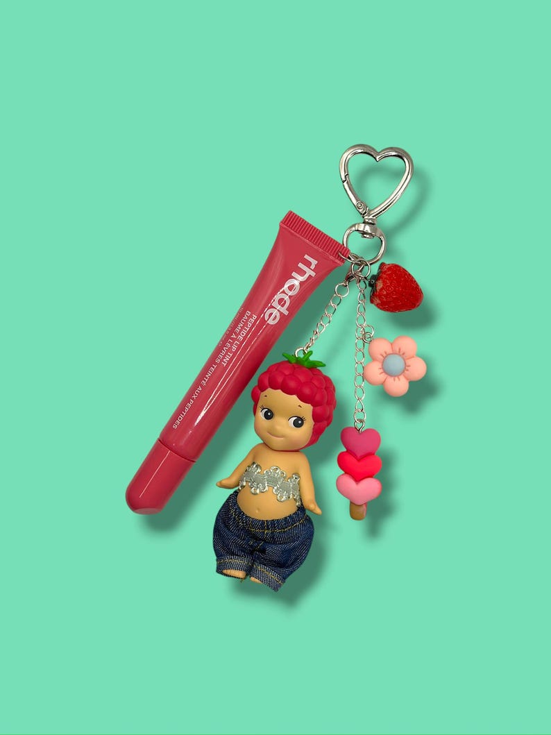 May include: A pink lip tint tube with the brand name "rhode" and a small doll keychain. The doll has a strawberry-shaped head, denim shorts, and a silver top. The keychain also includes a strawberry, flower, and heart charms.
