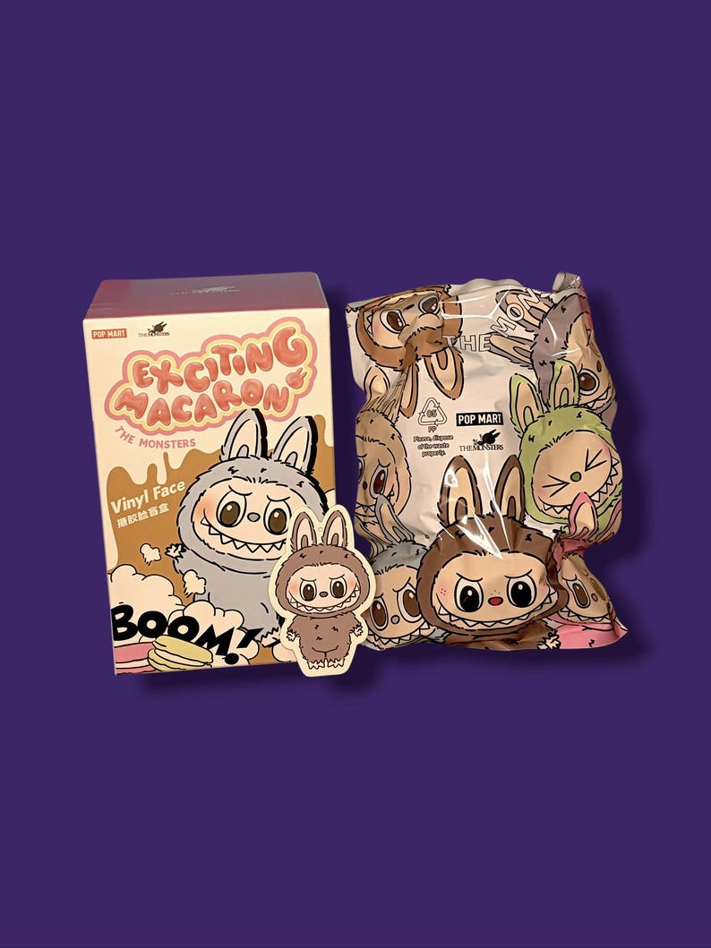 K&ouml;nnte beinhalten: Eine Pop Mart-Box und ein versiegelter Beutel mit The Monsters "Exciting Macaron" Vinyl-Gesichtsfiguren. Die Box ist beige mit rosa Text und einem Cartoon-Hasen-Design. Der Beutel zeigt verschiedene hasenartige Charaktere.