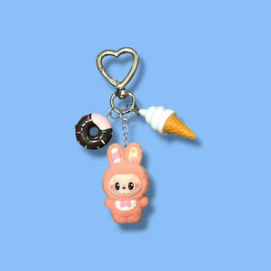 May include: A keychain with a heart-shaped clasp, featuring a donut, ice cream cone, and a pink bunny character. The donut is brown with pink frosting and sprinkles. The ice cream cone has a white swirl and a yellow cone. The bunny is pink.