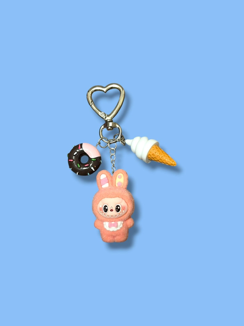 May include: A keychain with a heart-shaped clasp, featuring a donut, ice cream cone, and a pink bunny character. The donut is brown with pink frosting and sprinkles. The ice cream cone has a white swirl and a yellow cone. The bunny is pink.