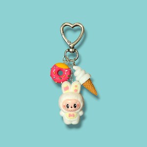 May include: A silver-colored keychain with a heart-shaped clasp. The keychain features three charms: a pink donut with yellow frosting, a white ice cream cone, and a white bunny figure with a pink bow. The background is a solid light blue.