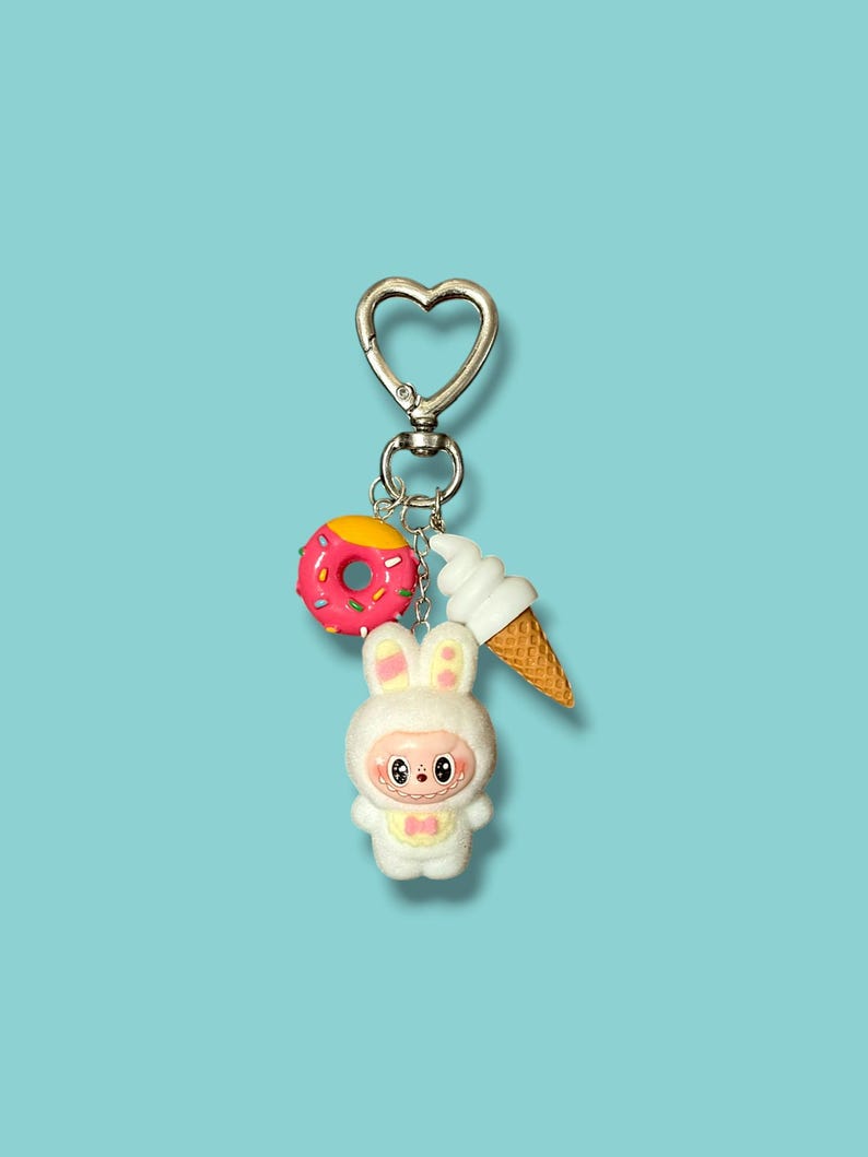 May include: A silver-colored keychain with a heart-shaped clasp. The keychain features three charms: a pink donut with yellow frosting, a white ice cream cone, and a white bunny figure with a pink bow. The background is a solid light blue.