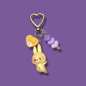 May include: A whimsical keychain featuring a gold-colored heart-shaped clasp. The keychain includes a yellow bunny charm, a yellow heart with the word "Love", and a purple ice cream cone charm. The background is a solid purple color.