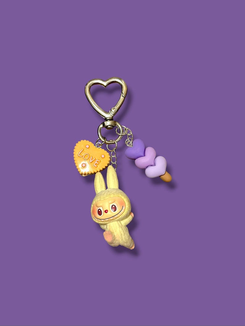 May include: A whimsical keychain featuring a gold-colored heart-shaped clasp. The keychain includes a yellow bunny charm, a yellow heart with the word "Love", and a purple ice cream cone charm. The background is a solid purple color.