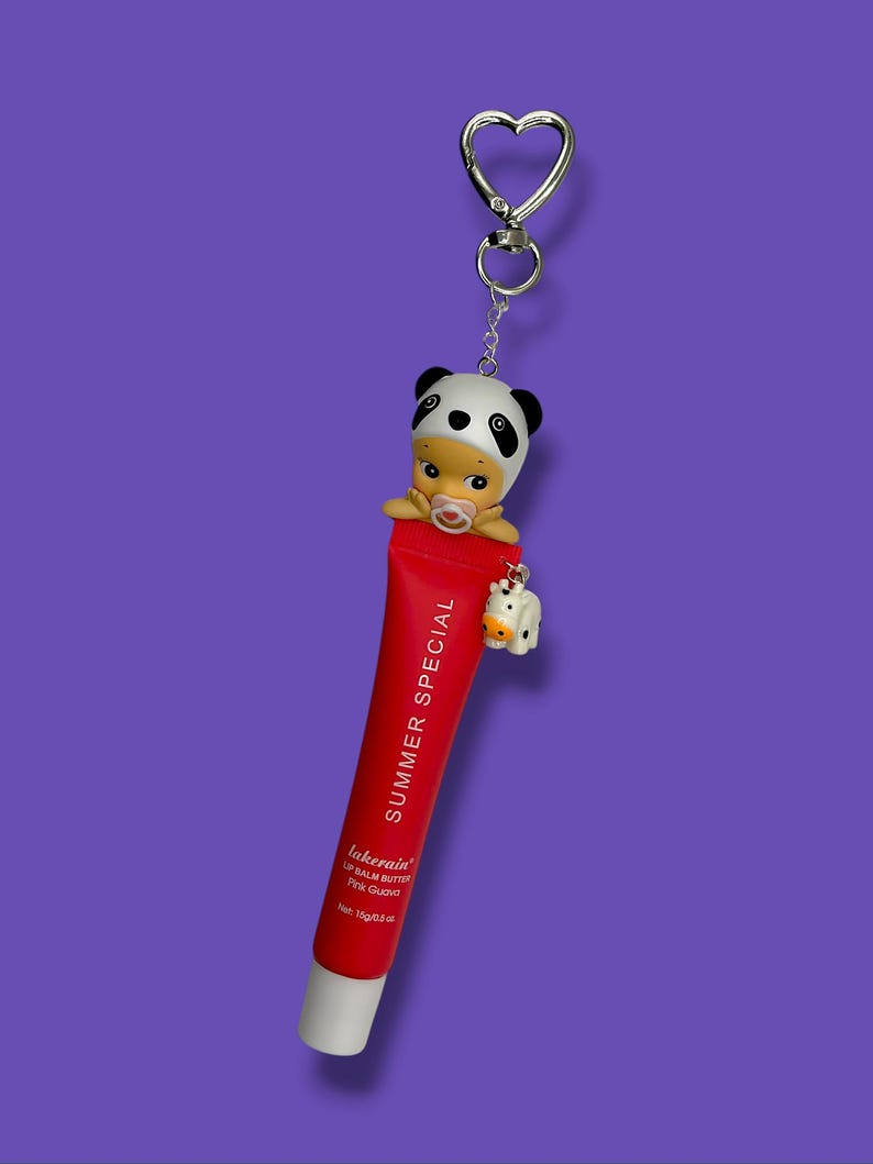 Op de afbeelding: Een rode lippenbalsem tube met de tekst "SUMMER SPECIAL" en "lakerain" erop, met een panda-thema bedel en een kleine koeienbedel. De lippenbalsem tube is bevestigd aan een zilveren hartvormige sleutelhanger.