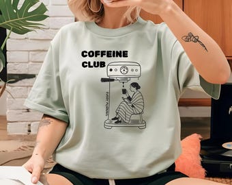 May include: A pale green t-shirt featuring the text "COFFEINE CLUB" above a black illustration of a coffee machine and a person. The words "EVERY MORNING" are printed vertically on the side of the machine.