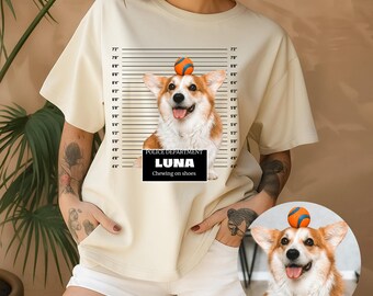 Op de afbeelding: Crèmekleurig T-shirt met een corgi-hond met een bal op zijn hoofd, tegen een achtergrond van een politie-opstelling. De naam van de hond, "LUNA", en de zin "Chewing on shoes" staan eronder. Het shirt is een casual kledingstuk.