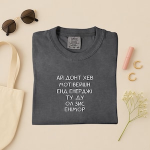 May include: A dark gray Comfort Colors t-shirt with white text that reads "I don't have motivation and energy to do all this anymore." The shirt is laid flat with accessories including sunglasses, a tote bag, and jewelry.