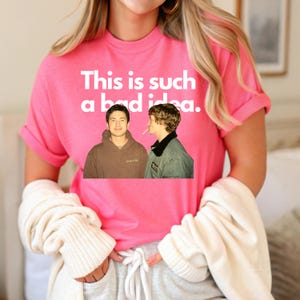May include: A pink t-shirt with the phrase "This is such a bad idea" in white text. The shirt features a graphic of two men. The person in the image is wearing a white sweater and gray sweatpants.