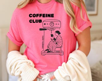 May include: A vibrant pink t-shirt featuring the text "COFFEINE CLUB" above a black illustration of a coffee machine and a person. The words "EVERY MORNING" are printed vertically on the side of the machine.