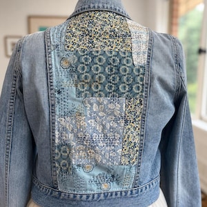 May include: Pale blue denim jacket with a patchwork design on the back. The patchwork features floral and geometric patterns in shades of blue, white, and yellow. The jacket is displayed on a mannequin.