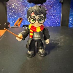 May include: A 3D-printed figurine of a character with black hair, round glasses, and a red and yellow scarf. The figure is wearing a black coat and holding a wand. The figurine is a collectible item.