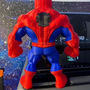 K&ouml;nnte beinhalten: Eine rot, blau und schwarz gehaltene Spider-Man-Actionfigur, von hinten betrachtet. Die Figur hat einen muskul&ouml;sen K&ouml;rperbau, mit roten Armen, Beinen und Torso, blauen Akzenten und einem schwarzen Spinnenemblem auf dem R&uuml;cken. Die Figur steht auf einer grauen Oberfl&auml;che.