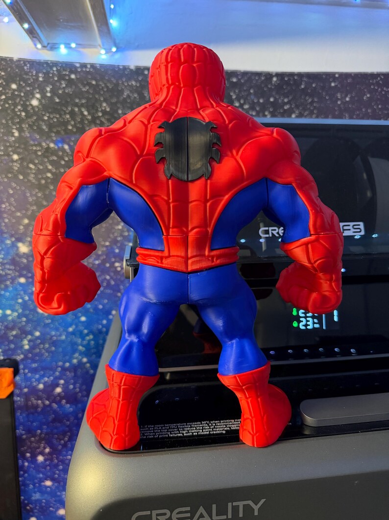 K&ouml;nnte beinhalten: Eine rot, blau und schwarz gehaltene Spider-Man-Actionfigur, von hinten betrachtet. Die Figur hat einen muskul&ouml;sen K&ouml;rperbau, mit roten Armen, Beinen und Torso, blauen Akzenten und einem schwarzen Spinnenemblem auf dem R&uuml;cken. Die Figur steht auf einer grauen Oberfl&auml;che.